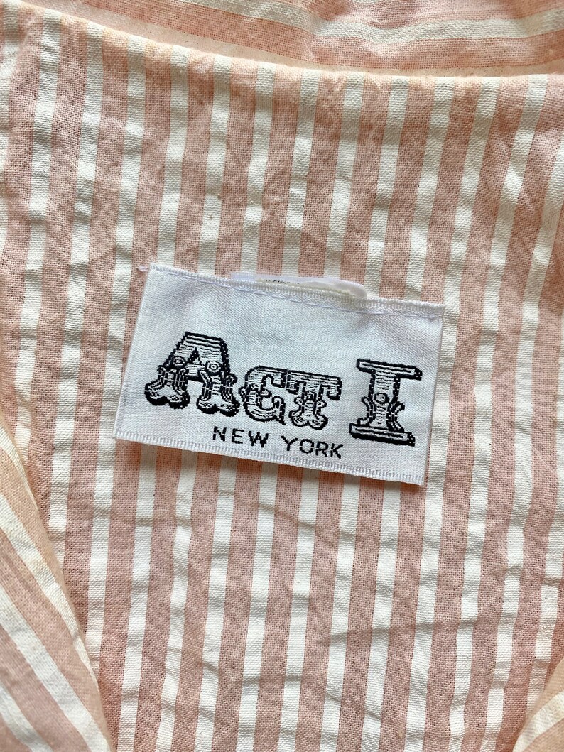 Pu&ograve; includere: Un'etichetta bianca con testo nero che recita "Act I NEW YORK" su un tessuto a righe rosa e bianche.