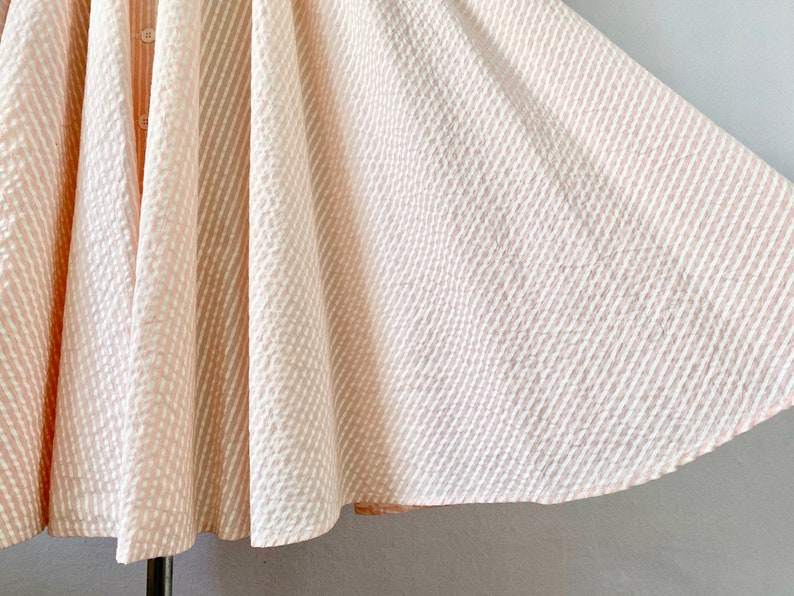 Pu&ograve; includere: Una gonna a righe rosa e bianche con una silhouette ampia e fluida. La gonna &egrave; realizzata in un tessuto leggero e ha una chiusura con bottone sul davanti.