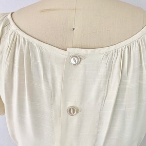 May include: A white button-up blouse with a gathered neckline and short sleeves. The blouse has a button closure down the back.