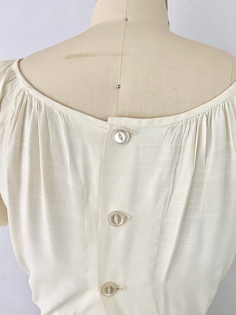 May include: A white button-up blouse with a gathered neckline and short sleeves. The blouse has a button closure down the back.