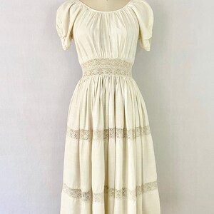 May include: A white cotton dress with a lace trim around the waist and hem. The dress has a scoop neck and short puff sleeves.