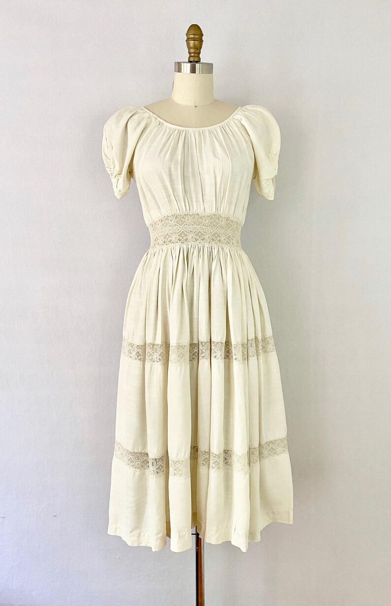 May include: A white cotton dress with a lace trim around the waist and hem. The dress has a scoop neck and short puff sleeves.