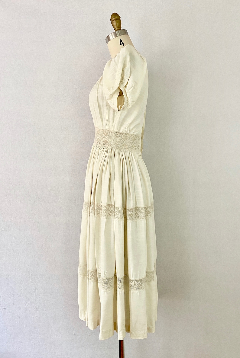 May include: A white cotton dress with a lace trim around the waist and hem. The dress has a fitted bodice and a full skirt.
