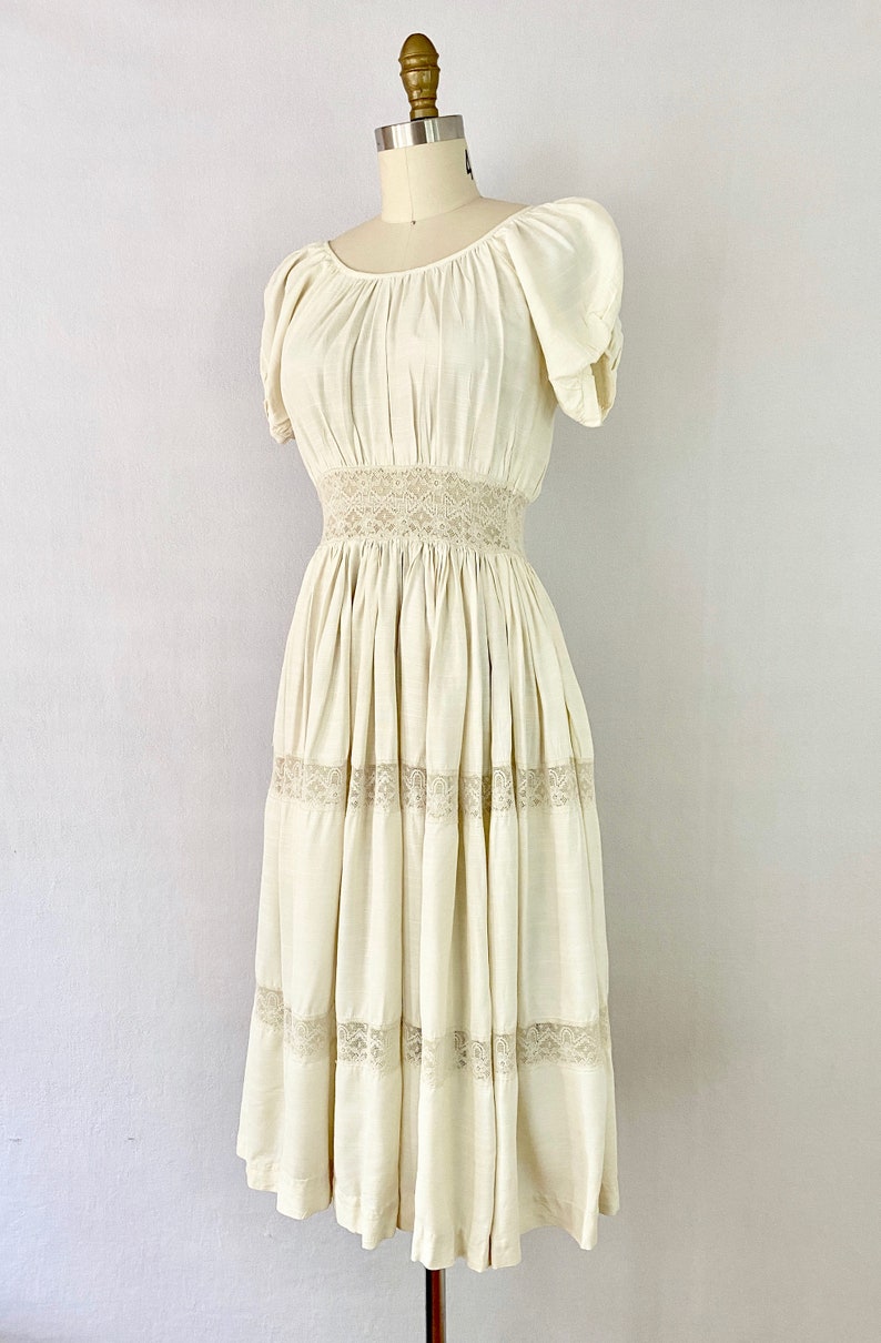 May include: A white vintage dress with a lace trim and a gathered skirt. The dress has a scoop neck and short puff sleeves.