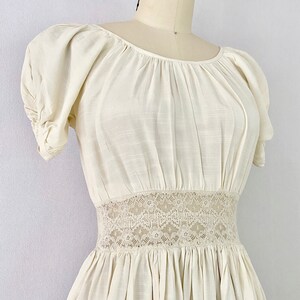 May include: A white cotton dress with a lace waistband and puffed sleeves. The dress has a gathered skirt and a scoop neckline.