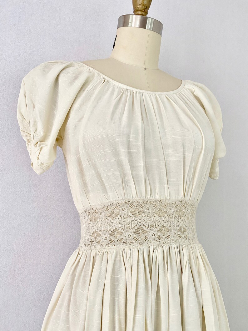 May include: A white cotton dress with a lace waistband and puffed sleeves. The dress has a gathered skirt and a scoop neckline.