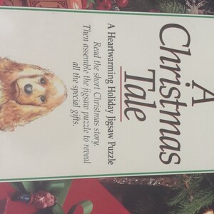 K&ouml;nnte beinhalten: Ein Weihnachts-Puzzle mit dem Titel "A Christmas Tale" mit einer Illustration eines braunen und wei&szlig;en Hundes. Die Puzzlebox hat einen gr&uuml;n-roten Hintergrund mit einem wei&szlig;en Rand.