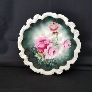 May include: A white porcelain bowl with a scalloped edge and a green and white floral design featuring pink roses. The bowl has a gold trim around the edge.
