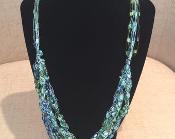 May include: A multi-strand necklace in shades of blue and green, displayed on a black jewelry stand. The necklace features woven strands with small beads, creating a textured and layered effect. The color palette includes various shades of blue and green.