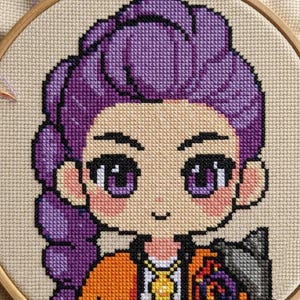 May include: Cross-stitch embroidery of a cartoon character with purple hair, large purple eyes, and an orange jacket. The embroidery is set in a wooden hoop, showcasing the detailed needlework and colour palette.