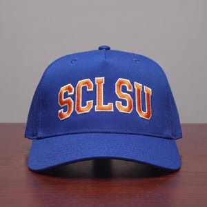 May include: A royal blue baseball cap featuring the letters SCLSU embroidered in orange with a white outline across the front panel. The cap has a structured crown, a curved brim, and a button on top, showcasing a classic five-panel design.