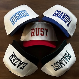 May include: Five white baseball caps with colored brims and embroidered text. The caps feature the words HUGHES in light blue, GRANDPA in dark blue, RUST in red, EAZER in black, and PSALMIST in dark green, all in a bold, block-style font.