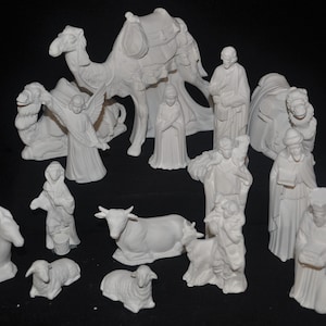 May include: A collection of unpainted white ceramic nativity figurines including a camel, an angel, a cow, sheep, and various human figures in robes. The set features detailed molded textures on each piece, presented as a complete group of decorative statues.