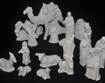 Könnte beinhalten: Eine Sammlung unbemalter weißer Keramikfiguren für eine Krippe, darunter ein Kamel, ein Engel, eine Kuh, Schafe und verschiedene menschliche Figuren in Gewändern. Das Set zeichnet sich durch detaillierte, geformte Texturen auf jeder Statue aus.
