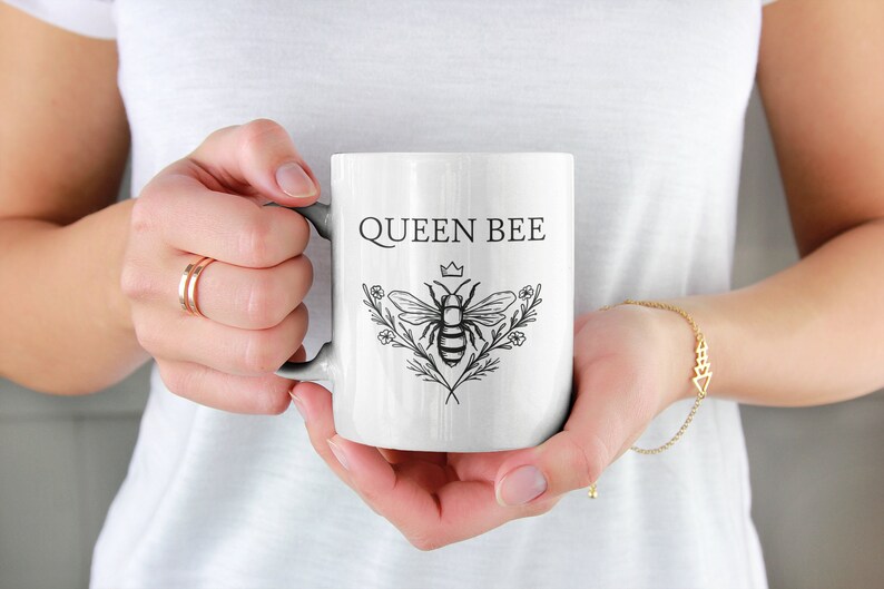May include: White ceramic mug with a black and white illustration of a bee with a crown and the text "QUEEN BEE".