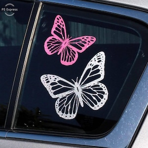 May include: Two butterfly decals on a car window. The top butterfly is pink with black accents. The bottom butterfly is white with black accents.