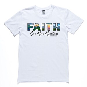 May include: White t-shirt with a graphic design that says "FAITH Can Move Mountains" with a mountain scene in the letters. The scripture reference "Matthew 17:20" is printed below the graphic.