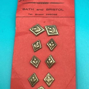 May include: A red rectangular card labeled Jessie's Button Box, Bath and Bristol, with the telephone number 299065. The card holds nine diamond-shaped metallic buttons, each featuring an embossed thistle design in a muted gold tone.