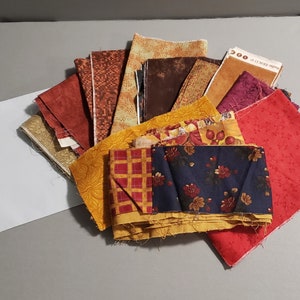 May include: A collection of fabric scraps in various autumnal colors and patterns, including red, orange, yellow, brown, and blue. The fabrics are arranged in a circular pattern, with the center of the circle being a piece of fabric with a floral design.