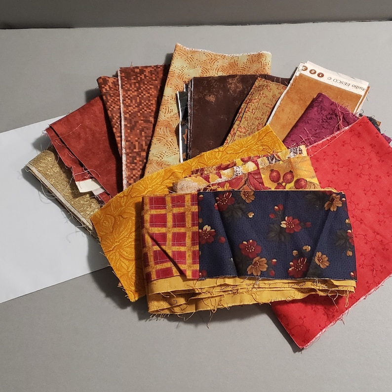 May include: A collection of fabric scraps in various autumnal colors and patterns, including red, orange, yellow, brown, and blue. The fabrics are arranged in a circular pattern, with the center of the circle being a piece of fabric with a floral design.