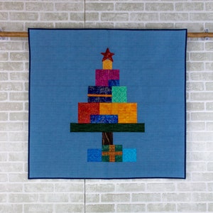 May include: A blue quilted wall hanging with a Christmas tree design made of colorful fabric squares. The tree is decorated with a red star and has a brown trunk.