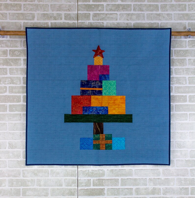 May include: A blue quilted wall hanging with a Christmas tree design made of colorful fabric squares. The tree is decorated with a red star and has a brown trunk.