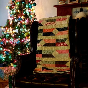 May include: A patchwork quilt with a geometric pattern in shades of pink, green, and brown is draped over a brown leather armchair. A Christmas tree with colorful ornaments and lights is in the background.
