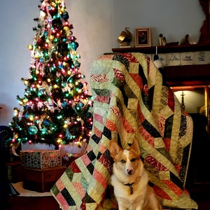 May include: A white and brown corgi dog sits under a colorful patchwork quilt in front of a decorated Christmas tree. The quilt has a variety of colors and patterns, including green, pink, black, and white.