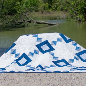 May include: A white and blue quilt with a geometric pattern is folded and placed on a gravel surface near a body of water.