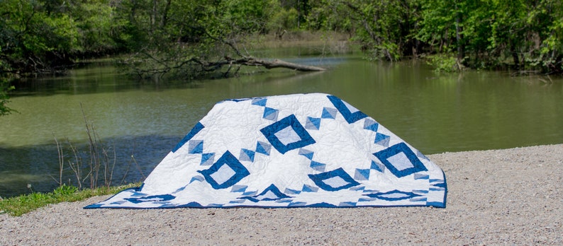 May include: A white and blue quilt with a geometric pattern is folded and placed on a gravel surface near a body of water.