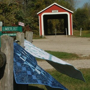 May include: A red covered bridge with white trim and the text "Celtun Bridge 1860" on the front. Two blue and white quilts are hanging on a wooden fence post in the foreground.
