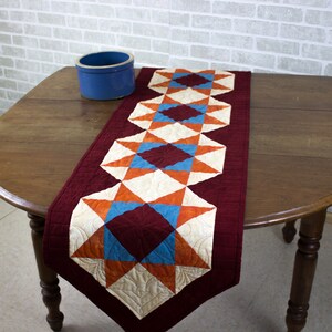 May include: A quilted table runner with a burgundy background and a pattern of orange, white, and blue stars and diamonds. The runner is on a wooden table.