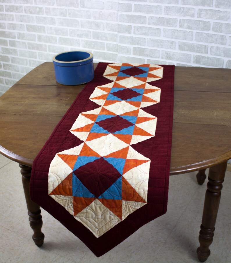 May include: A quilted table runner with a burgundy background and a pattern of orange, white, and blue stars and diamonds. The runner is on a wooden table.