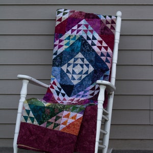 May include: A white rocking chair with a colorful patchwork quilt draped over it. The quilt is made of various shades of blue, green, red, and purple.