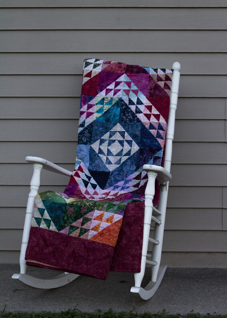 May include: A white rocking chair with a colorful patchwork quilt draped over it. The quilt is made of various shades of blue, green, red, and purple.