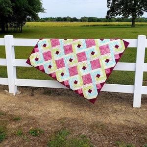 May include: A quilt with a geometric pattern in shades of pink, green, and blue is draped over a white wooden fence. The quilt is made of fabric squares sewn together to create a diamond pattern. The quilt is hanging in a rural setting with a field of sunflowers in the background.