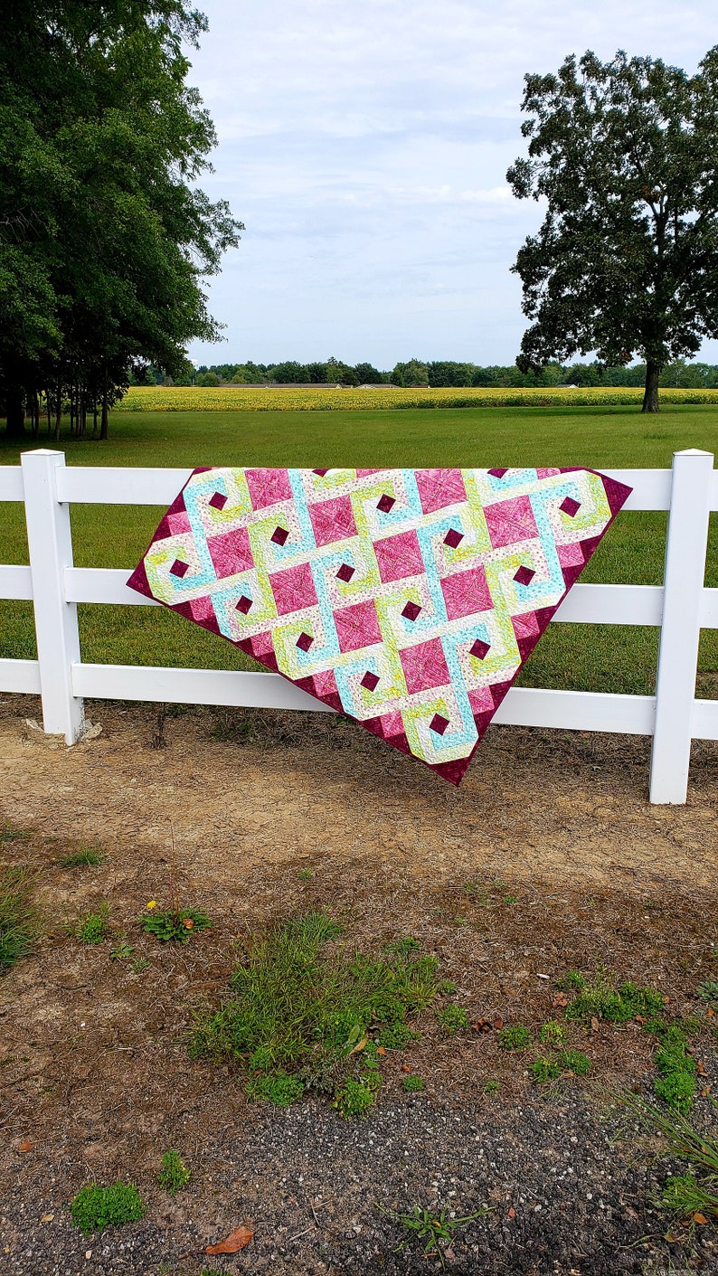 May include: A quilt with a geometric pattern in shades of pink, green, and blue is draped over a white wooden fence. The quilt is made of fabric squares sewn together to create a diamond pattern. The quilt is hanging in a rural setting with a field of sunflowers in the background.