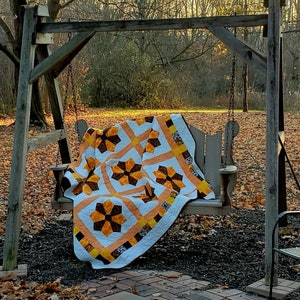 Op de afbeelding: Een wit, oranje en bruin patchwork-deken met een bloemenpatroon is over een houten schommel in een achtertuin met gevallen bladeren gelegd.