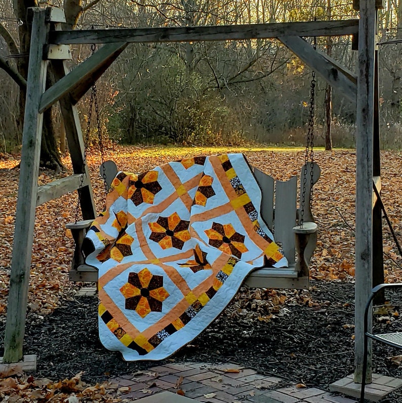 Op de afbeelding: Een wit, oranje en bruin patchwork-deken met een bloemenpatroon is over een houten schommel in een achtertuin met gevallen bladeren gelegd.