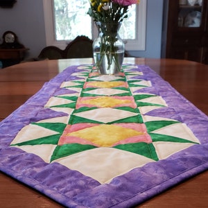 May include: A purple table runner with a geometric pattern of green, pink, and yellow triangles. The runner is on a wooden table with a vase of flowers in the background.