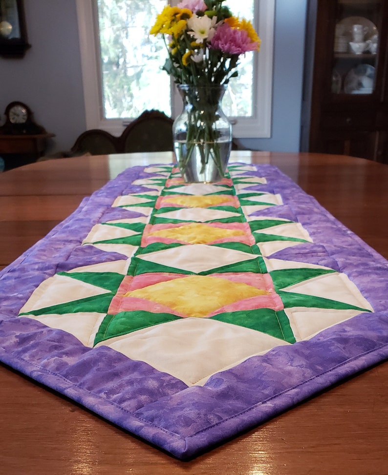 May include: A purple table runner with a geometric pattern of green, pink, and yellow triangles. The runner is on a wooden table with a vase of flowers in the background.