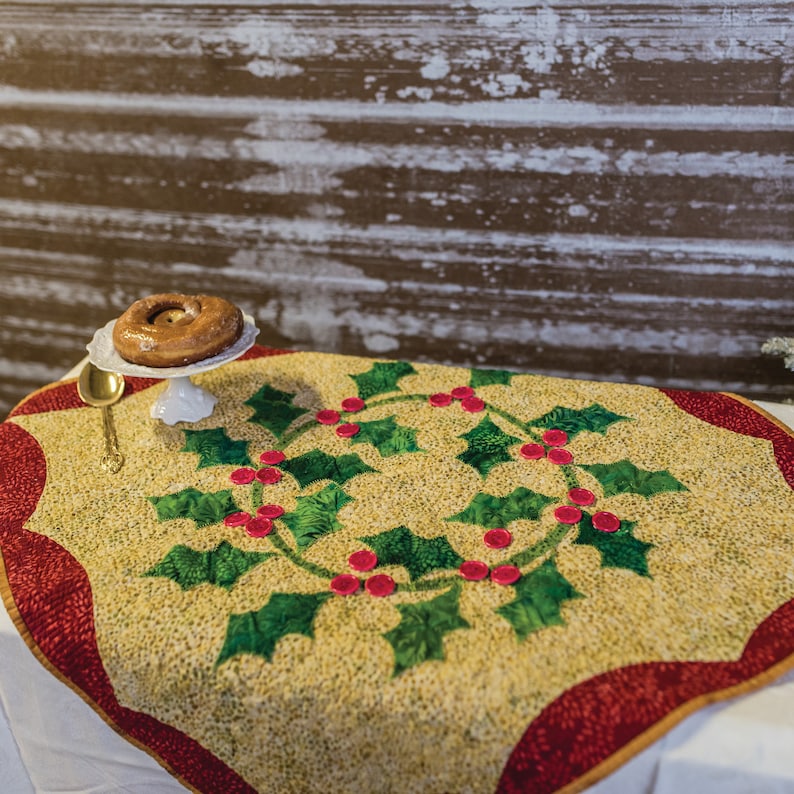 May include: A gold and red table runner with a green holly leaf and red berry design. The table runner is on a white table with a white cake stand and a donut on top.