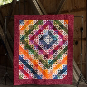 May include: A colorful quilt with a geometric pattern of triangles and diamonds. The quilt is displayed on a stand in front of a wooden structure.