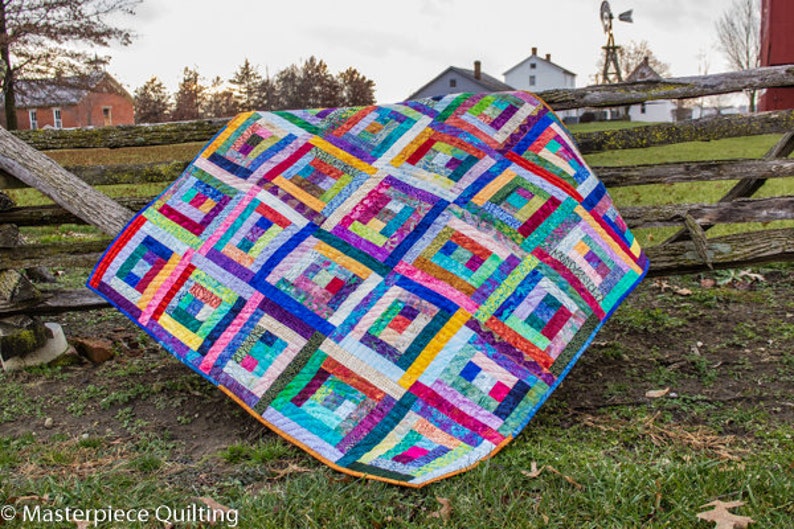 K&ouml;nnte beinhalten: Ein farbenfroher Quilt mit einem Blockhausmuster, der eine Vielzahl von leuchtenden Farben und Stoffen zeigt. Der Quilt ist &uuml;ber einen Holzzaun gelegt.