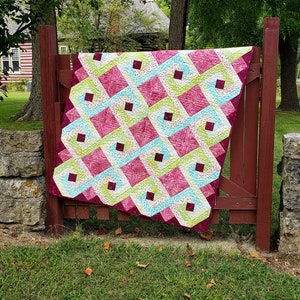 May include: A colorful quilt with a geometric pattern in shades of pink, green, and white. The quilt is hanging on a wooden fence in a backyard setting.