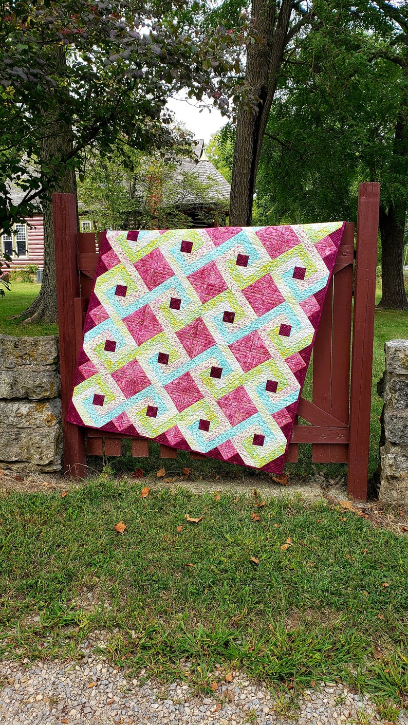 May include: A colorful quilt with a geometric pattern in shades of pink, green, and white. The quilt is hanging on a wooden fence in a backyard setting.