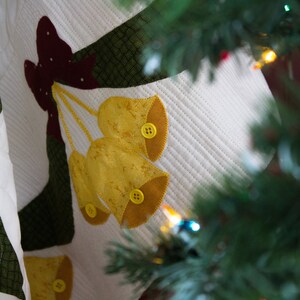 May include: A white quilted fabric with a green and burgundy applique of a bow and three yellow bells with yellow buttons. The fabric is partially obscured by a green pine branch.