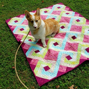 May include: A corgi dog sits on a pink, blue, and green quilt with a diamond pattern. The quilt is on a green grassy lawn.
