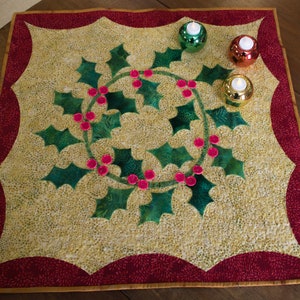 May include: A quilted table runner with a green holly leaf design and red berries. The runner has a red border with a gold trim. There are three small candle holders with white candles on the runner.