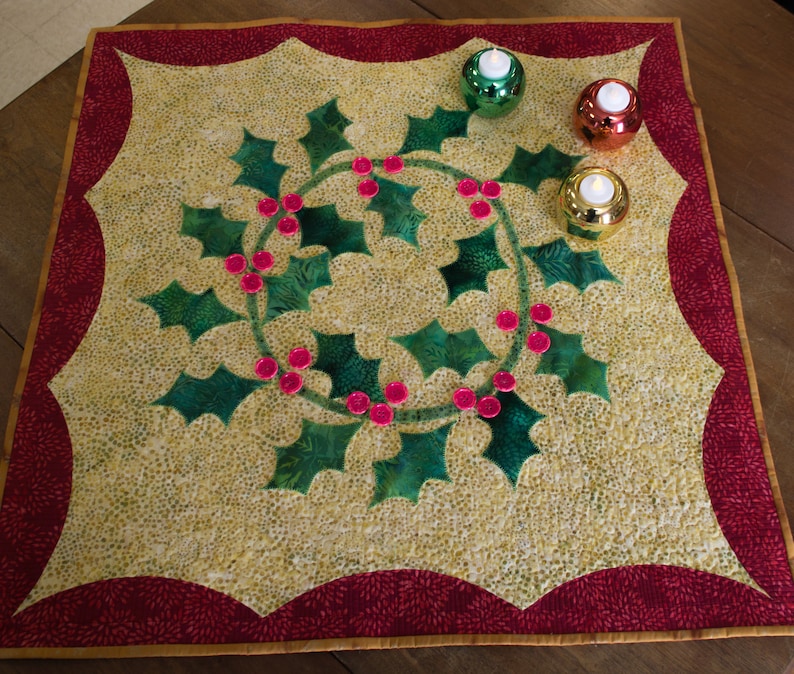 May include: A quilted table runner with a green holly leaf design and red berries. The runner has a red border with a gold trim. There are three small candle holders with white candles on the runner.